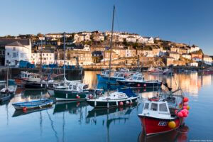 Pink dawn sky above Mevagissey village. A historic fishing harbour with a curving sheltering harbour wall. Flat calm sea. Boats moored. Houses on the hillside. South Cornish coast.