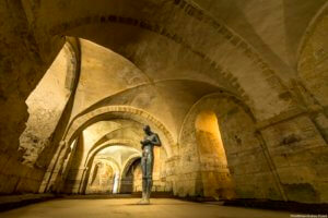 The underground crypt of Winchester cathedral, and a statue by Antony Gormley, called "Sound II", installed in 1986, and a modern shrine to Saint Swithun. A figure looking down at cupped hands.